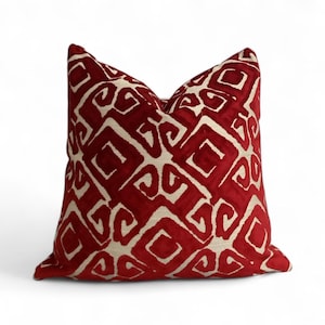 Pode incluir: Uma almofada decorativa quadrada com fundo vermelho e um padrão geométrico branco sujo repetido. O padrão consiste em quadrados e linhas curvas. A almofada tem uma aparência macia e texturizada.