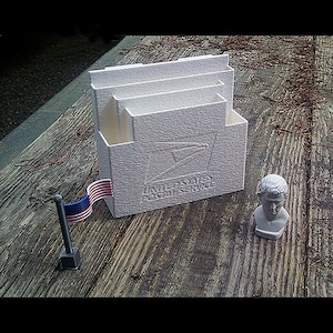 May include: A white textured desk organiser with the United States Postal Service logo and text. A small American flag and a miniature bust of a man are also present on a wooden surface.
