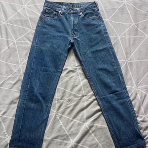 Può includere: Jeans in denim blu con chiusura a bottoni, design a cinque tasche e gamba dritta. I jeans sono di un lavaggio medio con sbiaditure e segni di usura sottili, che suggeriscono un'estetica vintage o usurata. I jeans sono stesi.