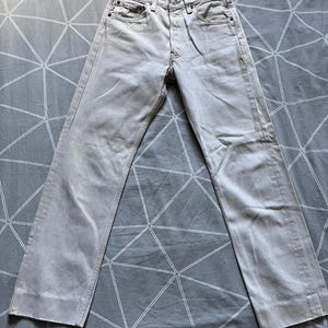 Può includere: Jeans in denim grigio chiaro con chiusura a bottoni, design a cinque tasche e gamba dritta. I jeans sono appoggiati su una superficie grigia a fantasia. Il tessuto sembra essere un denim di peso medio.