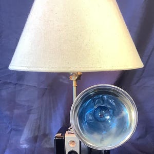 May include: A unique lamp featuring a vintage Brownie Hawkeye camera base. The black and silver camera is mounted on a wooden base, topped with a cream-colored lampshade. A large blue flashbulb is attached to the camera. The lamp is a creative blend of retro and functional design.