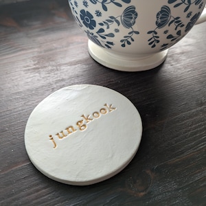 Op de afbeelding: Een ronde witte onderzetter met een getextureerd oppervlak en de naam jungkook in gouden reliëfletters. De onderzetter staat naast een witte keramische mok die is versierd met een blauw bloemenpatroon op de buitenkant.