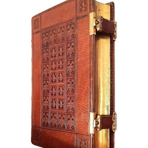 May include: A vintage, leather-bound book with an embossed fleur-de-lis design. The cover is a rich brown, with gold-gilded edges and decorative metal clasps. The book's spine is also gold, giving it a classic, antique appearance.