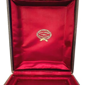 Peut inclure: Boîte ancienne ouverte, intérieur en velours rouge. La boîte est doublée de velours rouge et porte un emblème doré au centre. L'extérieur est en matériau foncé, peut-être en cuir. Un petit fermoir métallique est visible en haut.