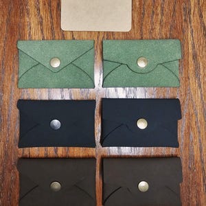 May include: A collection of handmade cardholders in various colors, including green, black, and brown. Each cardholder features a unique envelope-style design with a snap closure. A blank rectangular card is also present. The cardholders are arranged on a wooden surface.