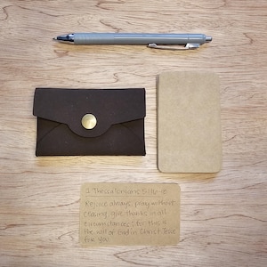 Puede incluir: Una cartera de cuero marrón oscuro con cierre de botón dorado, un pequeño bloc de notas beige y un bolígrafo gris están colocados sobre una superficie de madera. También hay una nota manuscrita con texto de 1 Tesalonicenses 5:16-18.
