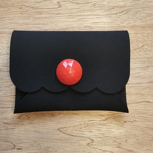 May include: A black felt clutch bag with a scalloped edge and a bright red, faceted button. The rectangular bag is closed. The background is a light brown wooden surface.
