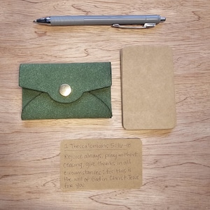 May include: An overhead shot of a green leather wallet with a gold button closure, a gray pen, a small tan notepad, and a handwritten note with a Bible verse. The items are arranged on a wooden surface.