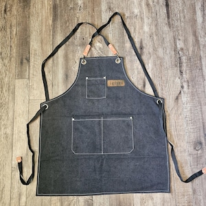 May include: A dark gray apron with two front pockets and a chest pocket. The apron has black straps and leather accents. The word "Tiphani" is on a small leather patch on the chest. The apron is made of a durable fabric.