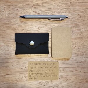 Könnte beinhalten: Ein schwarzer Lederkartenhalter mit silbernem Knopf, ein kleiner Notizblock und ein grauer Stift liegen auf einer Holzoberfläche. Eine handgeschriebene Notiz mit einem Bibelvers ist ebenfalls vorhanden. Die Artikel sind wahrscheinlich für den täglichen Gebrauch.