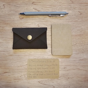 May include: A dark brown leather cardholder with a gold button closure, a gray pen, a small tan notepad, and a handwritten note with text from 1 Thessalonians 5:16-18. The items are arranged on a wooden surface.