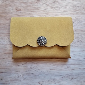May include: A small, mustard-yellow wallet with a scalloped flap and a decorative silver button closure. The wallet is made of a textured material and is closed. The background is a light wood grain.
