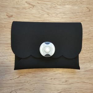 May include: A black clutch with a scalloped flap and a circular, white and gray clasp. The clutch is made of a soft material and is closed with a button clasp. The design is simple and elegant.