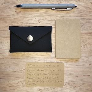 Könnte beinhalten: Ein schwarzer Kartenhalter mit silbernem Knopf, ein grauer Stift und eine leere beige Karte liegen auf einer Holzoberfläche. Eine handschriftliche Notiz mit Text aus 1. Thessalonicher 5:16-18 ist ebenfalls vorhanden.