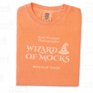 May include: An orange t-shirt with a crew neck, folded neatly. The shirt features the text "Real Product Photography" and "WIZARD OF MOCKS MOCKUP SHOP" in a stylized font, along with a wizard hat graphic. A tag is visible on the collar.