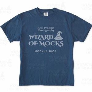 以下が含まれることがあります： 紺色のTシャツ。テキスト「Real Product Photography」と「WIZARD OF MOCKS MOCKUP SHOP」は薄いグレー。デザインには魔法使いの帽子グラフィックが含まれています。Tシャツはクルーネックです。