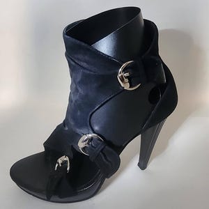 May include: A black high-heeled sandal boot featuring a peep-toe design, a tall stiletto heel, and three silver-toned metal buckles across the front and side. The upper is constructed from a soft, matte black material with cut-out details.