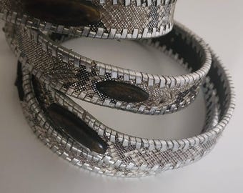 May include: Silver-toned metal bracelets with a snake-skin pattern and dark brown oval accents. The bracelets are stacked on top of each other, creating a layered effect. The design features a woven texture.