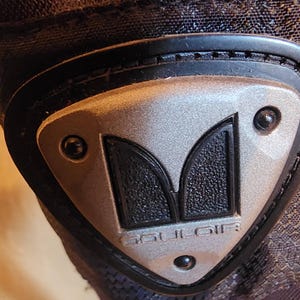 May include: A black fabric garment featuring a triangular silver-toned emblem with a black textured center. The emblem displays the stylized letter M and the text GOULOIR embossed in silver. It is secured with three small black rivets.