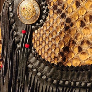 May include: Close-up of a black leather jacket with fringe and a snakeskin panel. The jacket features a decorative silver and gold-toned circular emblem with a red gem and red beads.