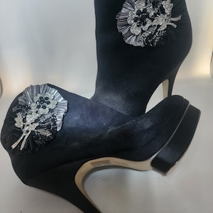 May include: A pair of black satin ankle boots with high stiletto heels and platforms. Each boot features a large, circular, pleated black fabric embellishment adorned with silver-tone metal floral accents and clear crystals.