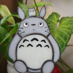 May include: A stained glass figure of Totoro, a character from the animated film My Neighbor Totoro. The figure is gray with a white belly and black details. It is held by two metal rods.