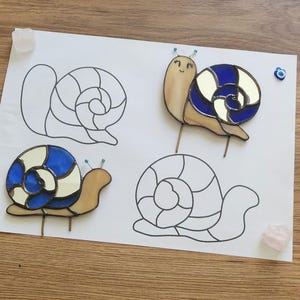 May include: Two stained glass snail figures featuring tan bodies and shells with alternating dark blue and cream-colored glass segments. The snails have two small antennae with blue beads at the tips. Two black-lined snail patterns are on the paper.