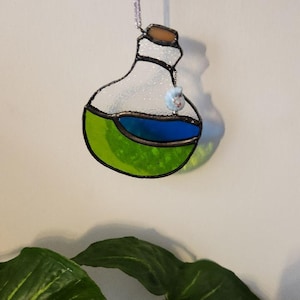 May include: Stained glass potion bottle ornament with green and blue liquid. The bottle has a brown cork and is outlined in black. A small charm hangs from the top. The ornament is suspended by a silver string.