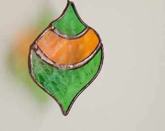May include: A stained glass ornament in a teardrop shape, featuring green and orange sections. The glass is framed with a dark metal and hangs from a chain. The ornament is suspended against a plain white background.