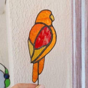 Pode incluir: Um ornamento de pássaro em vitral em tons de laranja, amarelo e vermelho, com contornos pretos. O pássaro é segurado por uma mão contra uma parede branca texturizada. O design do pássaro lembra um vitral.