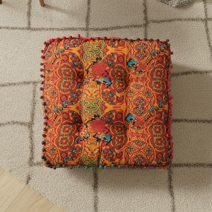 May include: Square floor cushion with a vibrant, ornate pattern in shades of orange, red, and blue. The cushion is trimmed with small, colourful pom-poms. The design features a buttoned centre.