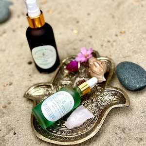 Puede incluir: Una bandeja dorada en forma de mano de Hamsa contiene una botella de aceite facial verde, un cristal de cuarzo rosa y una peque&ntilde;a flor rosa. Un frasco rociador negro con tapa dorada est&aacute; en el fondo. Los art&iacute;culos est&aacute;n sobre una superficie arenosa.