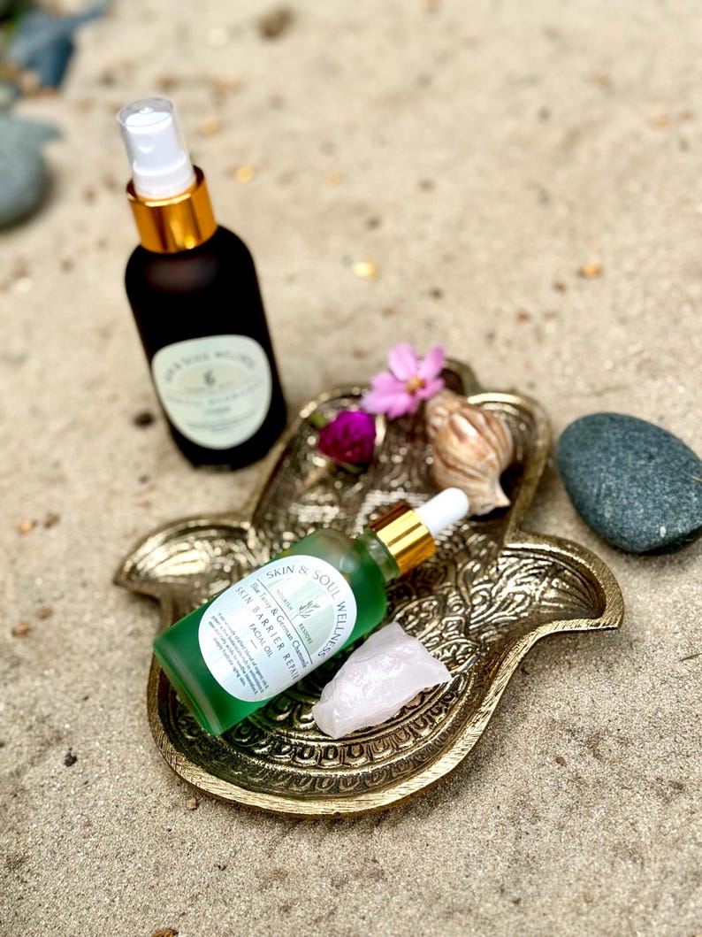 Puede incluir: Una bandeja dorada en forma de mano de Hamsa contiene una botella de aceite facial verde, un cristal de cuarzo rosa y una peque&ntilde;a flor rosa. Un frasco rociador negro con tapa dorada est&aacute; en el fondo. Los art&iacute;culos est&aacute;n sobre una superficie arenosa.
