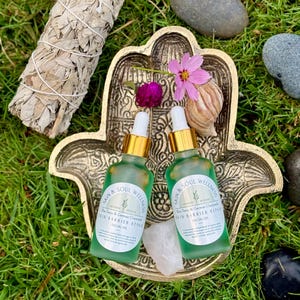 Puede incluir: Dos botellas de vidrio verde de aceite facial con cuentagotas dorados se encuentran en un plato dorado en forma de mano de Hamsa. Un manojo de salvia blanca, flores y piedras est&aacute;n dispuestos alrededor del plato sobre hierba verde. Las botellas tienen etiquetas que dicen "Skin & Soul Wellness".