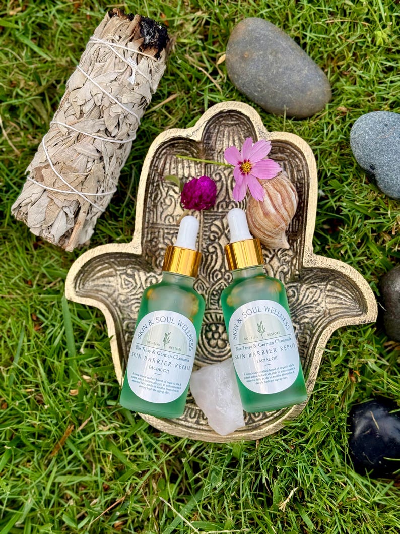 Puede incluir: Dos botellas de vidrio verde de aceite facial con cuentagotas dorados se encuentran en un plato dorado en forma de mano de Hamsa. Un manojo de salvia blanca, flores y piedras est&aacute;n dispuestos alrededor del plato sobre hierba verde. Las botellas tienen etiquetas que dicen "Skin & Soul Wellness".