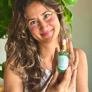 Puede incluir: Una mujer sostiene una peque&ntilde;a botella de vidrio de s&eacute;rum para el cuidado de la piel. La botella tiene una tapa dorada y un l&iacute;quido verde en su interior, con una etiqueta blanca que dice "Earth & Soul Wellness". La mujer est&aacute; sonriendo.