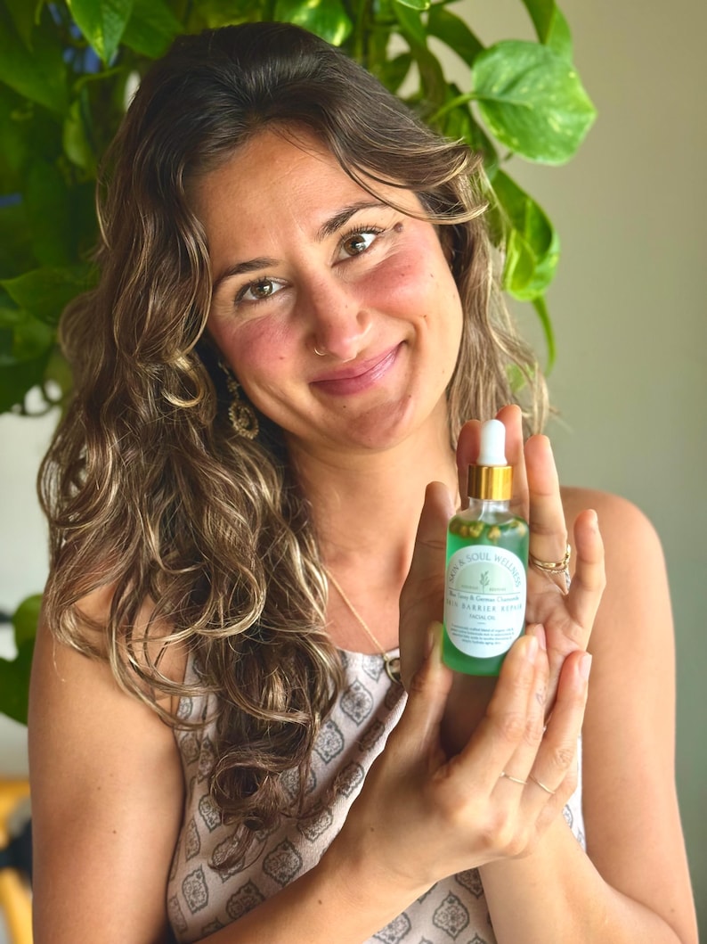 Puede incluir: Una mujer sostiene una peque&ntilde;a botella de vidrio de s&eacute;rum para el cuidado de la piel. La botella tiene una tapa dorada y un l&iacute;quido verde en su interior, con una etiqueta blanca que dice "Earth & Soul Wellness". La mujer est&aacute; sonriendo.