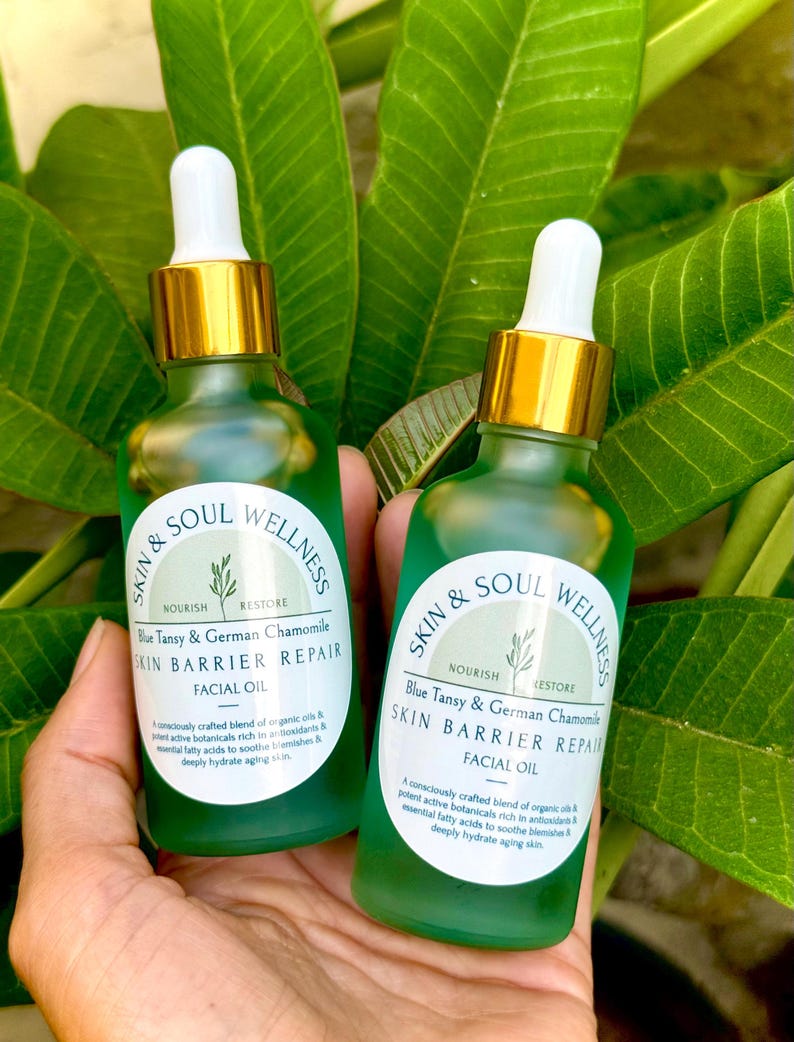 Puede incluir: Dos botellas de vidrio verde de aceite facial con cuentagotas dorados se sostienen frente a hojas verdes. Las botellas tienen etiquetas blancas con el texto "Skin & Soul Wellness" y "Blue Tansy & German Chamomile SKIN BARRIER REPAIR FACIAL OIL."