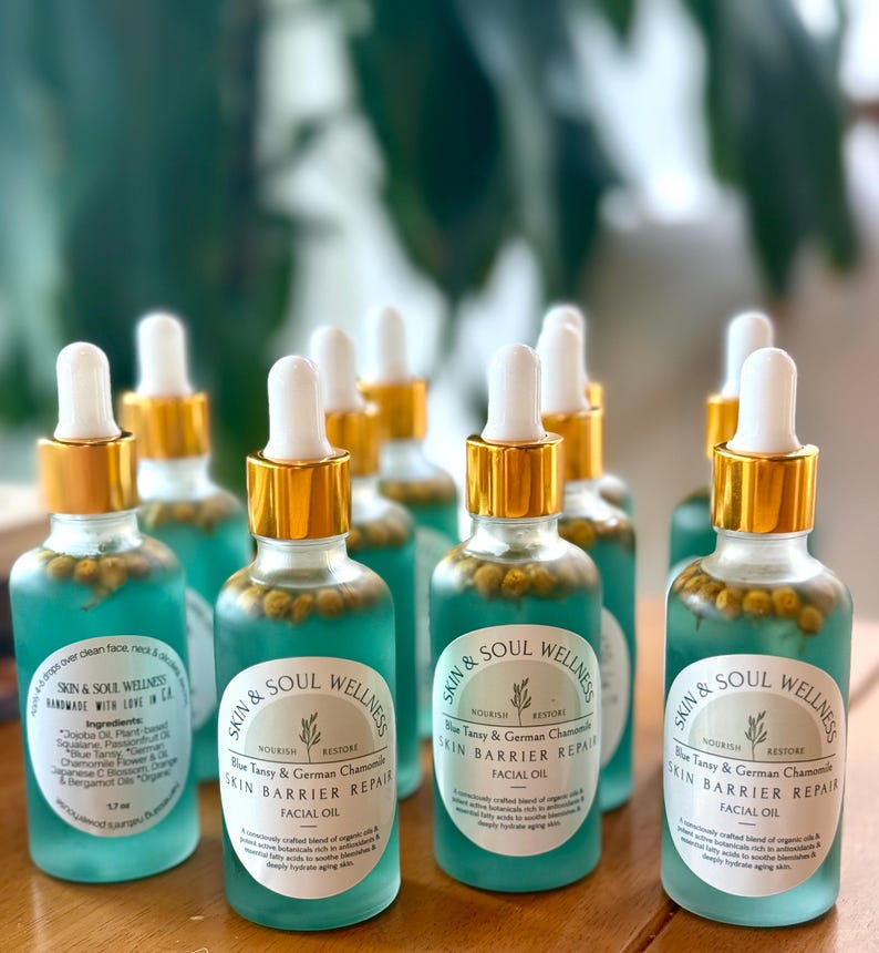 Puede incluir: Una colecci&oacute;n de botellas de aceite facial Skin & Soul Wellness. Cada botella de vidrio transparente tiene una tapa dorada y un cuentagotas blanco. El l&iacute;quido azul contiene peque&ntilde;os capullos de flores amarillas. La etiqueta dice "Skin Barrier Repair Facial Oil."