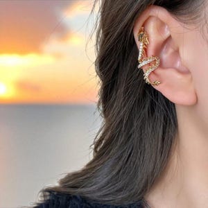 Può includere: Un orecchino a cerchio dorato a forma di drago, incrostato di piccoli cristalli trasparenti. Il drago si curva lungo l'orecchio, con la testa in alto e la coda in basso. Lo sfondo mostra un tramonto sull'oceano.