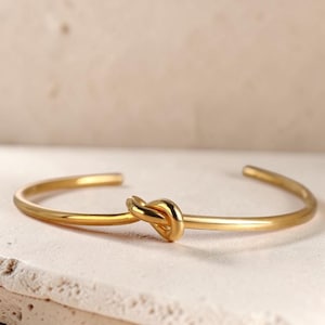 May include: A gold-colored cuff bracelet with a knot design. The bracelet is made of a shiny, smooth metal and has an open design. The knot is centered on the bracelet, adding a unique detail to the jewelry.