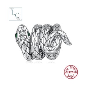 Pode incluir: Um anel de cobra em prata com padrão de escamas texturizado, duas pedras redondas transparentes na cabeça e duas pedras verdes nas laterais. O anel possui o selo 925 sterling silver. O logotipo LC é visível no canto superior esquerdo.