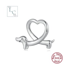 May include: A polished silver charm shaped like a dachshund dog with a long body that curves upward to form a heart outline. The dog features a small, clear, round gemstone set into its head. A circular stamp reads Real Sterling 925 Silver.