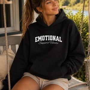 May include: Black hoodie with the text "EMOTIONAL Support Hoodie" in white block letters. The hoodie has a drawstring hood and a front pocket. The model is wearing the hoodie and tan shorts.