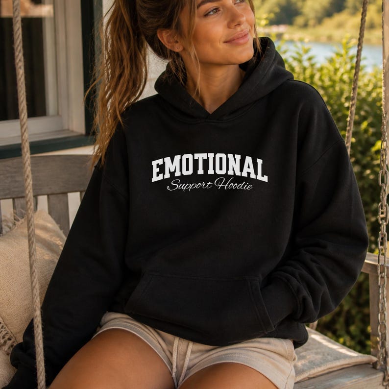 May include: Black hoodie with the text "EMOTIONAL Support Hoodie" in white block letters. The hoodie has a drawstring hood and a front pocket. The model is wearing the hoodie and tan shorts.