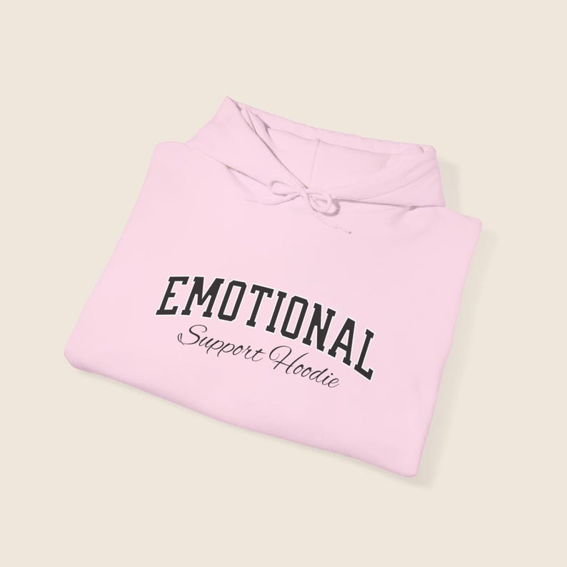 May include: A light pink hoodie with the words "EMOTIONAL" in a bold, black, arched font above "Support Hoodie" in a smaller, cursive font. The hoodie has a drawstring and a folded hood.