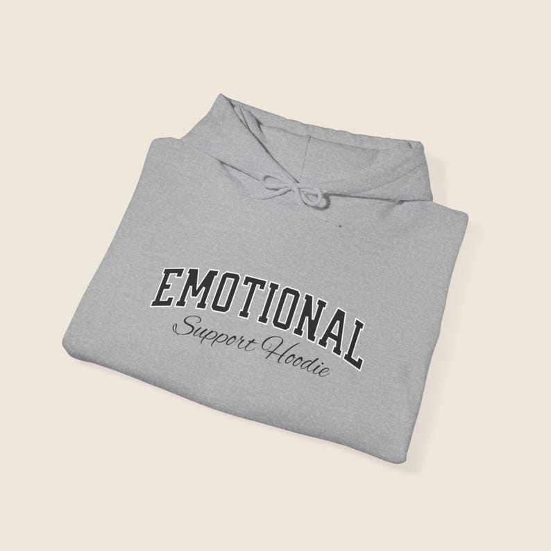 May include: A folded light gray hoodie with the words "EMOTIONAL" arched across the chest in a black, collegiate-style font. Below, in a script font, is "Support Hoodie". The hoodie has a drawstring hood.