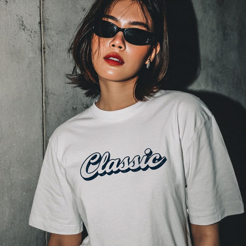 May include: White t-shirt with the word "Classic" in a navy blue script font. The shirt has short sleeves and a relaxed fit. The model is wearing black sunglasses.