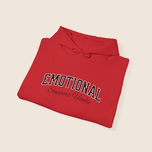 May include: A bright red hoodie with the text "EMOTIONAL" in a black and white collegiate font. Below the word "EMOTIONAL" is the text "Support Hoodie" in a cursive font. The hoodie is folded neatly.