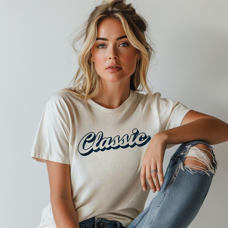 May include: Cream-colored t-shirt with the word "Classic" in navy blue script. The shirt is paired with light blue denim jeans with distressed details. The model is wearing a silver ring.